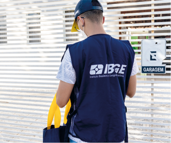 Fotografia de uma pessoa vistas de costas próximo ao portão de uma residência. Ela usa um boné azul, está com um colete azul com a logo do I B G E, e segura uma sacola.