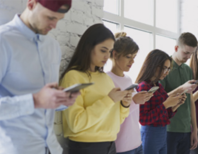 Foto de cinco jovens enfileirados, todos olhando atentamente para seus celulares. Eles estão em pé, próximos a uma parede branca de tijolos, com uma grande janela ao fundo que permite a entrada de luz natural. Cada pessoa segura o celular com as duas mãos, demonstrando concentração nas telas. O grupo é diverso, com homens e mulheres usando roupas casuais em diferentes cores, como amarelo, vermelho, azul e verde.