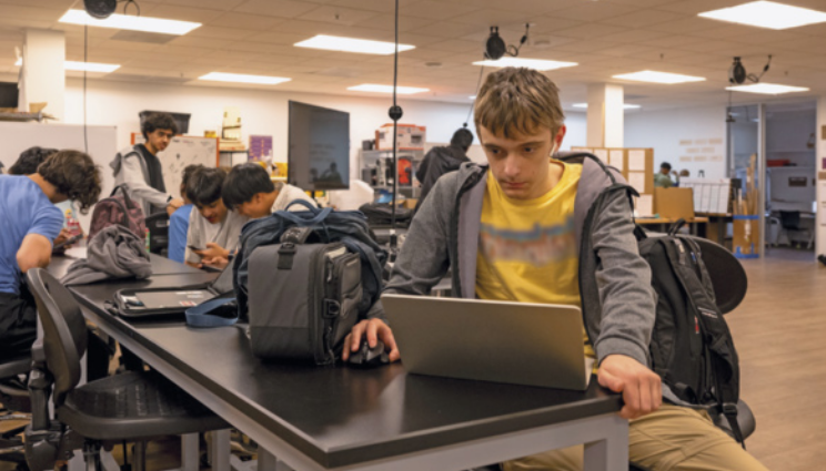 Fotografia de menino sentado e mexendo em um notebook, ele usa uma camisa amarela e casaco, ao seu lado há uma mochila. Ao fundo, há outas pessoas conversando e mexendo em celulares, há também prateleiras com caixas e uma televisão.