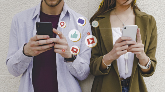 Fotografia de duas pessoas, um homem e uma mulher, vistas da cintura para cima, usando seus smartphones lado a lado. Em volta dos aparelhos, aparecem ícones digitais flutuantes representando redes sociais e interações online, como um coração, um polegar positivo, um balão de mensagem, um símbolo de localização e os logos do YouTube e do Instagram. A imagem simboliza o uso cotidiano das redes sociais, a comunicação digital e a constante conexão com plataformas online.