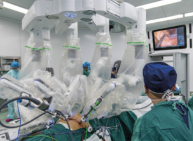 Imagem de uma sala de cirurgia equipada com um sistema robótico cirúrgico com múltiplos braços articulados cobertos por material esterilizado. Diversos profissionais de saúde em trajes cirúrgicos verdes estão posicionados ao redor do paciente, acompanhando o procedimento. Um monitor no canto superior direito exibe uma imagem ampliada do interior do corpo do paciente.