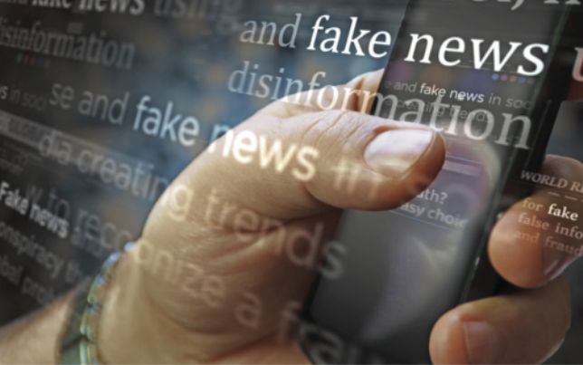 Foto de uma mão segurando um celular, com a tela refletindo luz. Sobre a imagem, há uma sobreposição de palavras como “fake news”, “disinformation” e outras expressões relacionadas à desinformação, escritas em diferentes tamanhos, posições e opacidades. As palavras aparecem em inglês e se destacam na composição, criando uma atmosfera de alerta sobre o conteúdo veiculado.