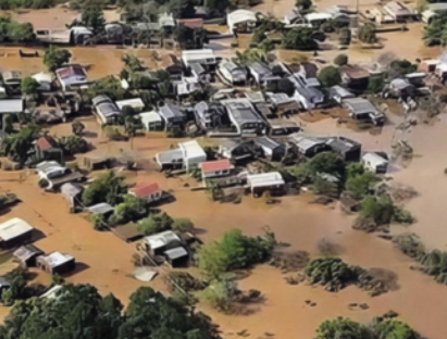 Fotografia aérea de uma área residencial afetada por uma enchente. A cena é dominada por uma grande extensão de água barrenta, de cor marrom, que cobre o solo e parte das estruturas das casas e das árvores.