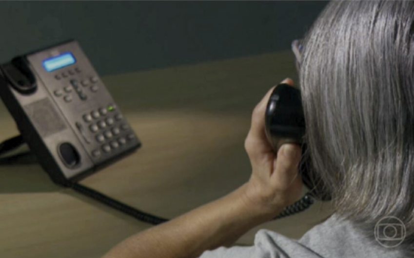 Fotografia mostrando uma pessoa idosa de cabelos grisalhos atendendo uma ligação em um telefone fixo. A imagem foca nas costas da pessoa e no aparelho telefônico sobre uma mesa. No canto inferior direito aparece discretamente o logotipo da TV Globo.