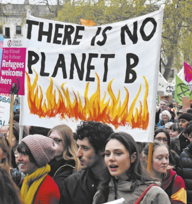 Foto de uma manifestação com várias pessoas reunidas em local aberto, algumas segurando cartazes, sendo o mais destacado um com a frase 'THERE IS NO PLANET B' escrita em letras maiúsculas pretas sobre fundo branco, com chamas pintadas abaixo do texto. Outros cartazes visíveis incluem dizeres como 'Refugees welcome here' e 'Say it loud say it clear'. Ao fundo, há árvores sem folhas e a fachada de um edifício antigo.