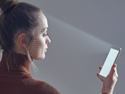 Fotografia de uma jovem mulher de perfil segurando um smartphone à sua frente, com projeções de luz e uma malha de pontos brancos sobre seu rosto, sugerindo o uso de tecnologia de reconhecimento facial; ela usa uma blusa de gola alta marrom e o fundo é neutro, desfocado e iluminado suavemente.
