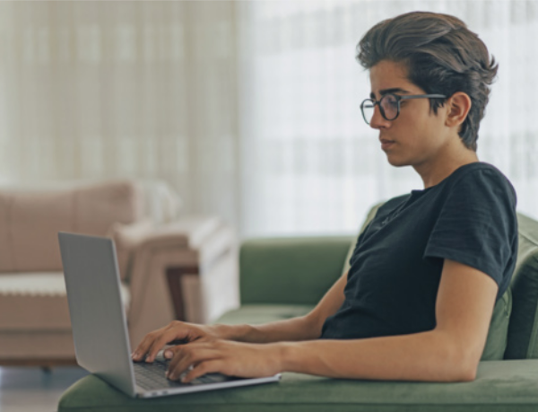 Foto de um jovem sentado em um sofá verde, utilizando um notebook no colo em um ambiente interno bem iluminado. Ele usa camiseta preta, usa óculos e mantém uma postura concentrada enquanto digita. Ao fundo, é possível ver uma cortina translúcida e parte de outro sofá.