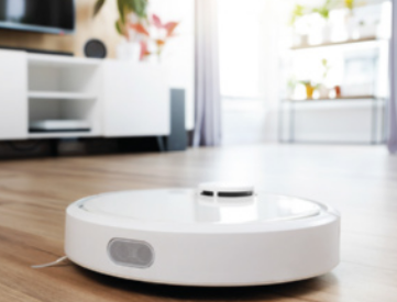 Imagem de um aspirador robô de formato circular e cor branca em funcionamento sobre um piso de madeira clara. O ambiente é uma sala de estar iluminada por luz natural, com móveis brancos ao fundo, televisão, plantas e prateleiras com objetos decorativos.