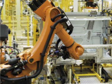 Imagem de um braço robótico industrial de cor laranja operando em uma linha de montagem automatizada. O braço está posicionado em frente à estrutura metálica de um veículo em construção. Ao fundo, é possível observar diversas máquinas, equipamentos e estruturas amarelas e cinzas, características de um ambiente de fábrica automatizada.
