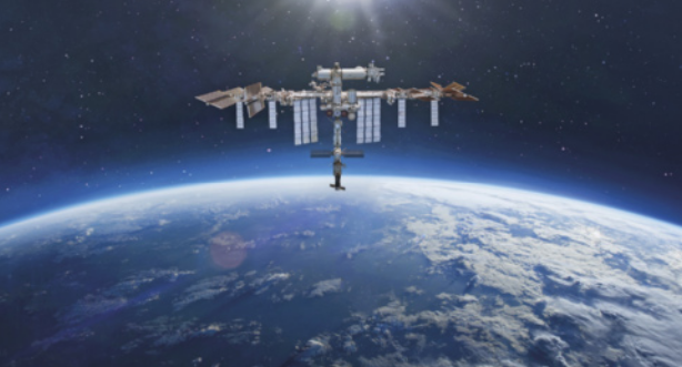 Imagem da Estação Espacial Internacional em órbita acima da Terra. A estação aparece centralizada na parte superior da imagem, com suas estruturas metálicas e painéis solares claramente visíveis. Ao fundo, o planeta Terra é visto parcialmente, com sua curvatura destacada e áreas cobertas por nuvens, oceanos e continentes sob a luz do Sol. O espaço ao redor é escuro, e o brilho do Sol aparece suavemente atrás da estação.