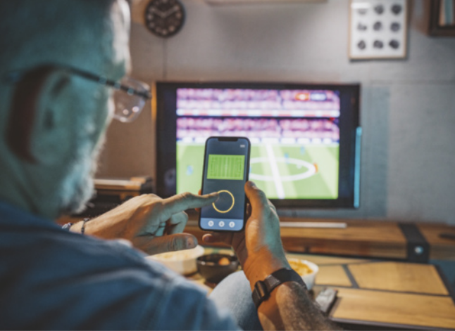 Foto de um homem de cabelos e barba grisalhos sentado em frente a uma televisão que exibe uma partida de futebol, segurando um celular com um aplicativo relacionado ao jogo, possivelmente de estatísticas ou interatividade, enquanto usa o dedo indicador da outra mão para tocar a tela do aparelho.