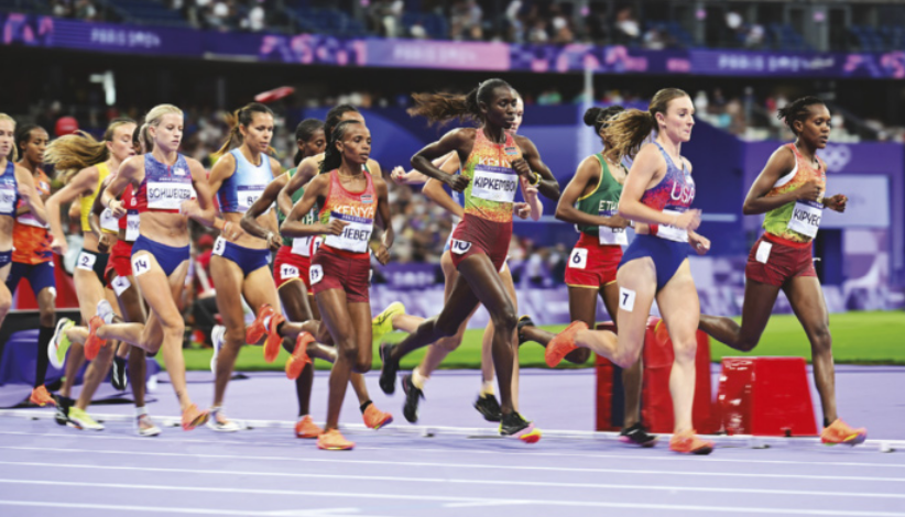 Fotografia de um grupo de  corredoras em uma corrida de resistência em um estádio, posicionadas muito próximas entre si.