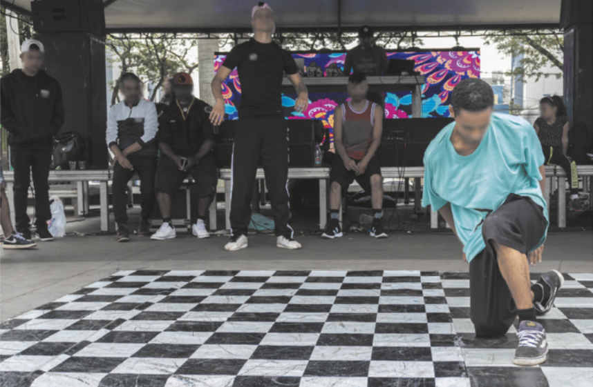 Fotografia de um homem dançando ajoelhado em uma pista de breaking, com um grupo de homens ao fundo, sentados e em pé, observando.