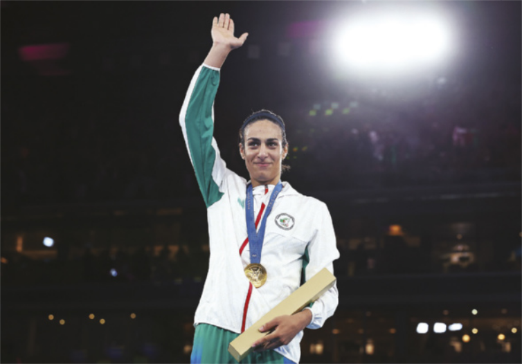 Fotografia de Imane Khelif. Ela é uma mulher com cabelos presos em um rabo-de-cavalo, usando uma medalha pendurada ao pescoço e segurando uma caixa contendo um diploma, com o braço erguido para o alto.
