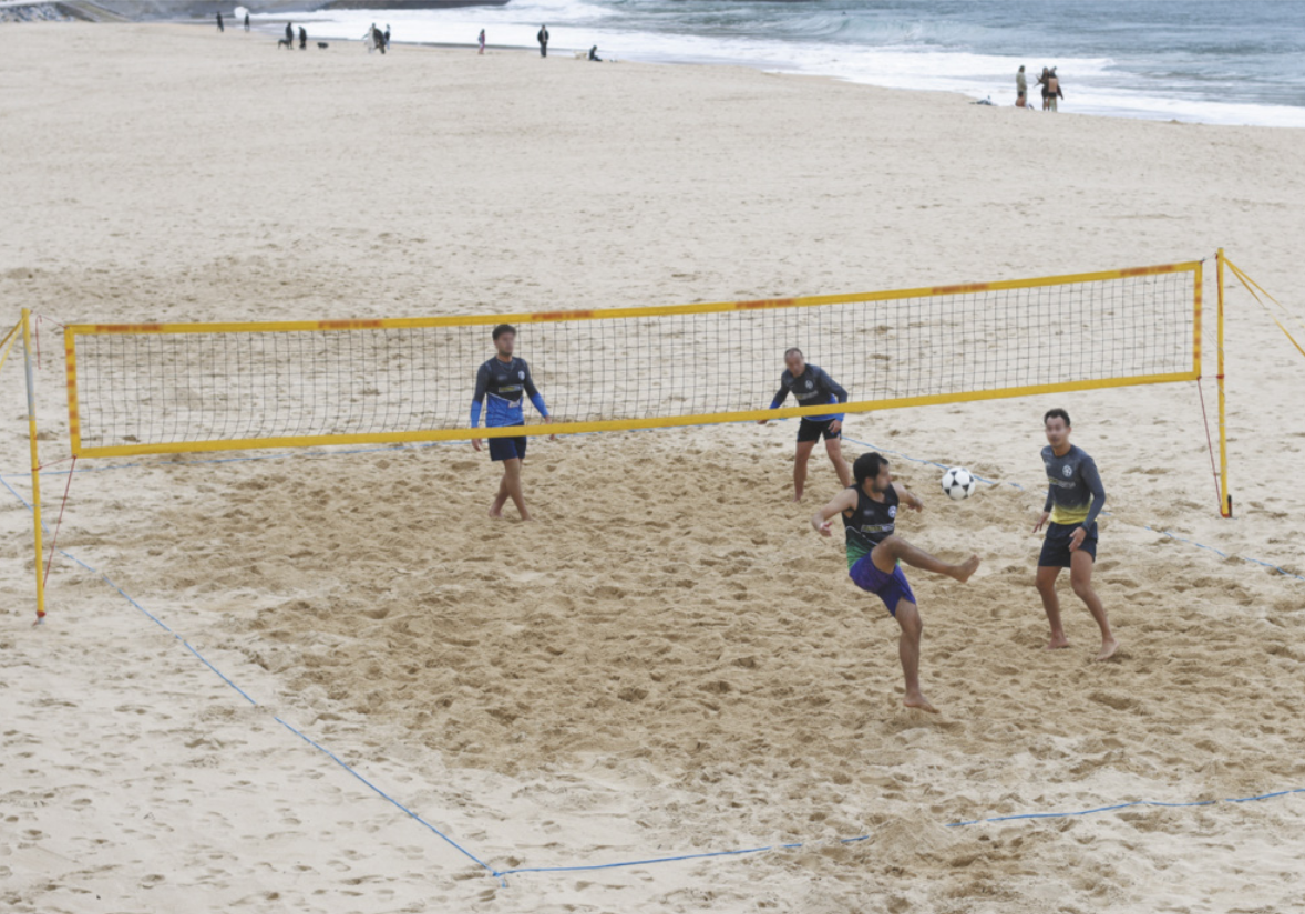 Fotografia de duas duplas de homens jogando futevôlei em uma quadra na areia da praia, com uma rede separando os dois lados da quadra. Um deles está de costas para a dupla adversária, chutando a bola em direção à rede com a lateral interna do pé.