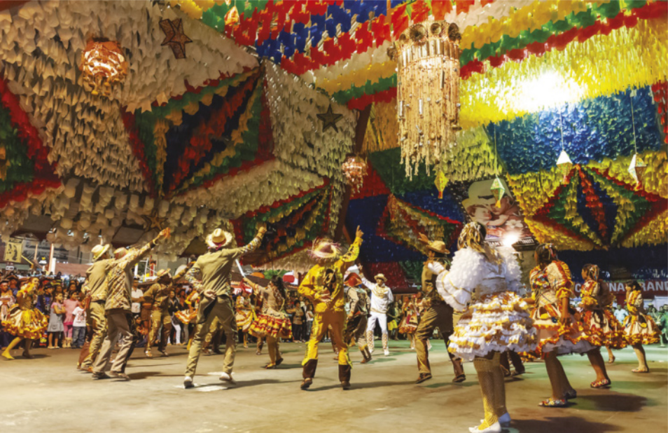 Fotografia de um grupo de pessoas dançando em coreografia em um salão amplo, enfeitado com bandeiras coloridas no teto. Os homens estão de chapéu, com um dos braços erguido, e as mulheres de vestido, com fitas e enfeites coloridos.