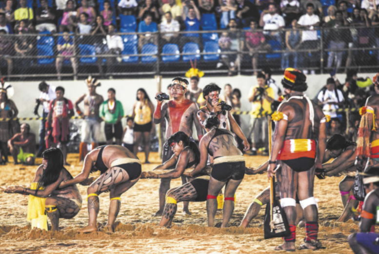 Fotografia de um grupo de mulheres indígenas com pinturas corporais e adereços tradicionais em fila, puxando um cabo de guerra em uma arena com piso de areia. Ao fundo há uma plateia em uma arquibancada, assistindo à competição.