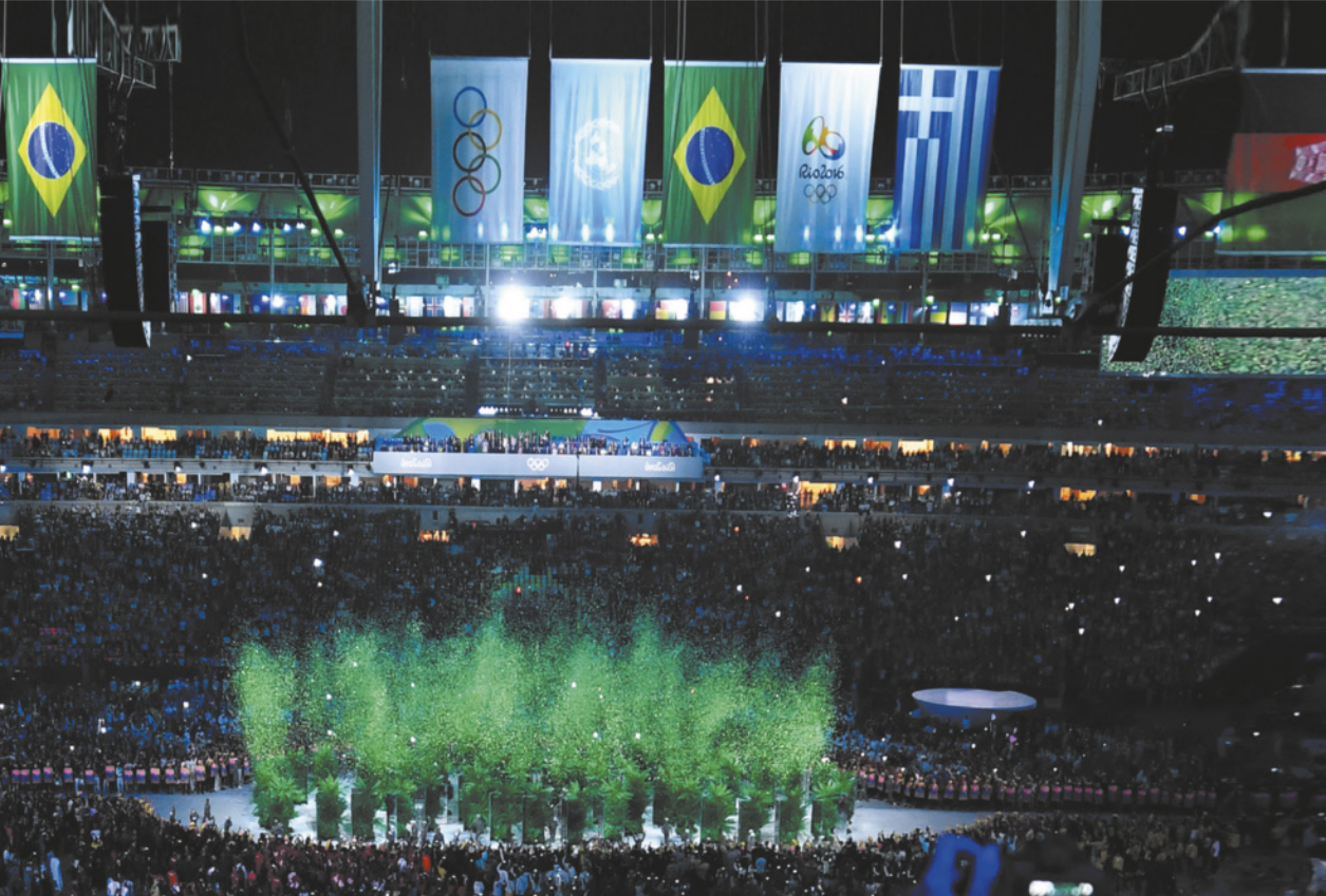 Fotografia de um espetáculo em um estádio, à noite, com a plateia em arquibancadas assistindo a uma apresentação no centro do estádio. No alto do estádio estão hasteadas as bandeiras do Brasil, da Grécia, dos Jogos Olímpicos, das Nações Unidas e da Olimpíada Rio 2016.