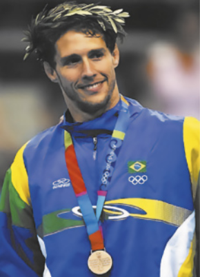Fotografia de Flávio Canto. Ele é um atleta com barba e cabelos curtos, usando uma coroa de louros e uma medalha pendurada ao pescoço, vestido com um uniforme com as cores da bandeira brasileira.