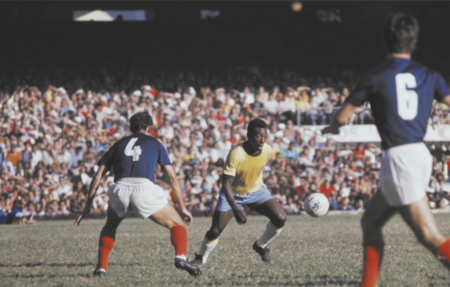 Fotografia de Pelé jogando futebol com o uniforme da seleção brasileira, no gramado de um estádio. Ele está controlando a bola, dando passadas com as pernas flexionadas, diante de dois jogadores adversários. Ao fundo está a torcida em uma arquibancada.