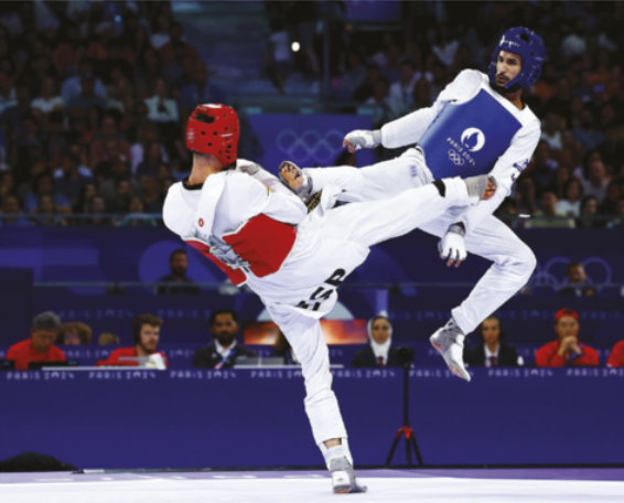 Fotografia de Edival Pontes durante uma luta de taekwondo olímpico. Ele está no ar, executando um chute que atinge o peito do adversário, que, por sua vez, se defende com o antebraço enquanto contra-ataca com um chute na região do quadril de Edival. Ambos os atletas vestem quimonos e utilizam equipamentos de proteção: capacetes, caneleiras, luvas e coletes acolchoados, um em azul e outro em vermelho.