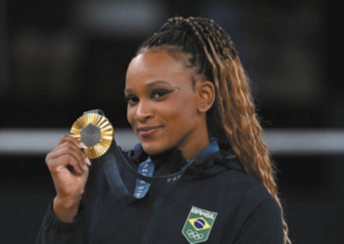 Fotografia de Rebeca Andrade. Ela é uma mulher de cabelos presos em tranças longas, usando maquiagem nos olhos, segurando uma medalha de ouro pendurada ao pescoço.