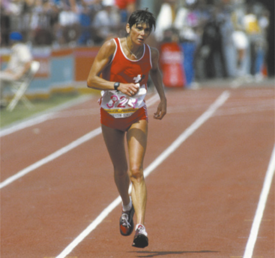 Fotografia de Gabriela Andersen-Schiess. Ela é uma mulher de cabelos curtos, correndo em uma pista de atletismo.