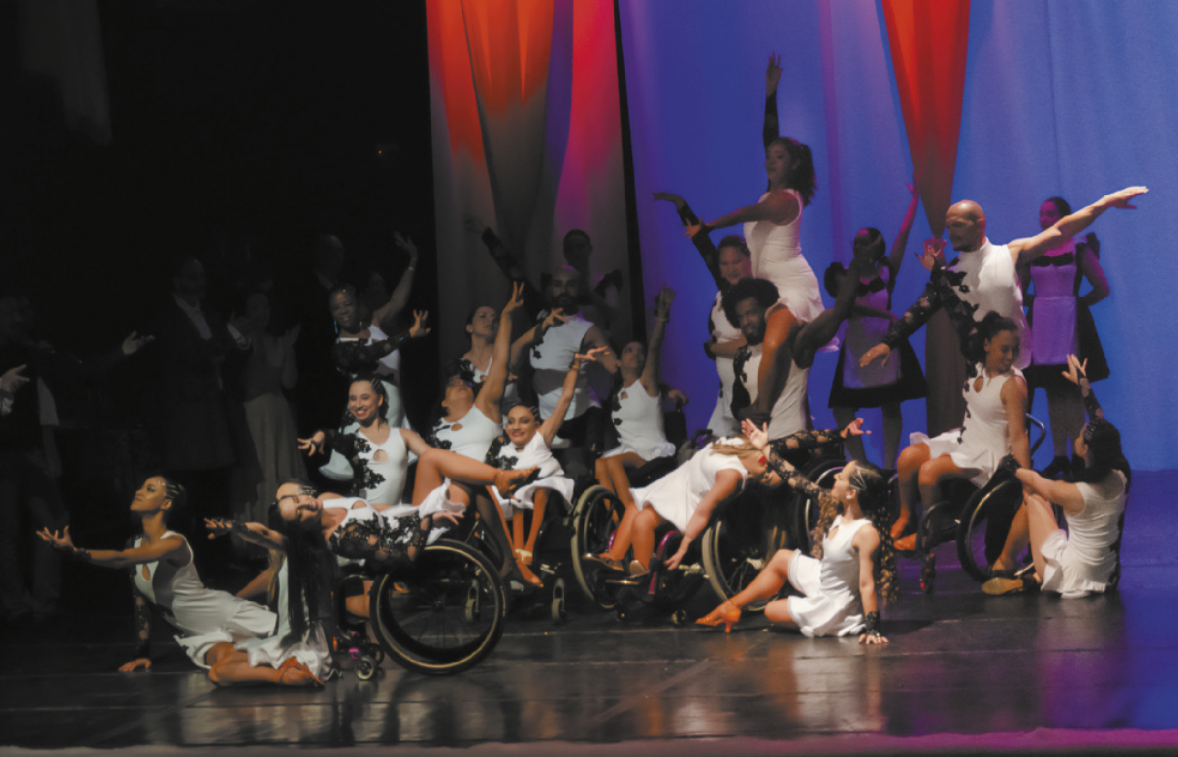 Fotografia de um grupo de bailarinas e bailarinos em um palco, algumas delas sentadas sobre cadeiras de roda, estendendo um dos braços para o alto.