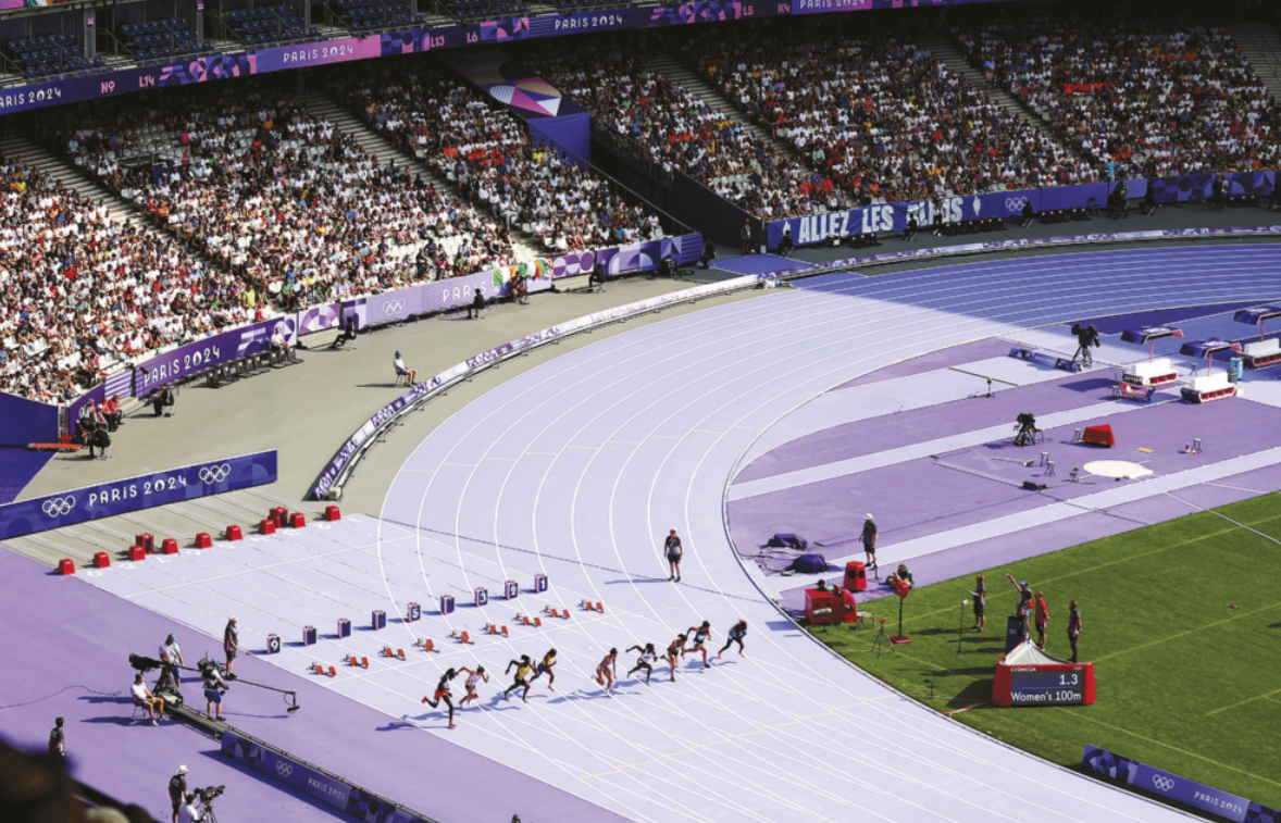 Fotografia de corredoras iniciando uma corrida em um estádio olímpico, com uma plateia assistindo a partir das arquibancadas. As corredoras estão correndo em alta velocidade, uma em cada raia da pista de atletismo.