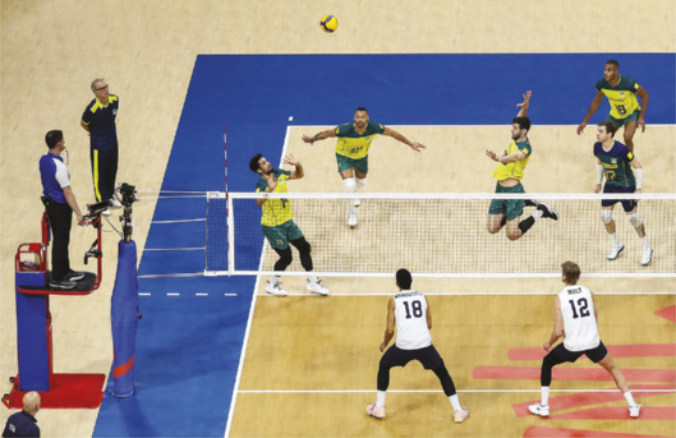 Fotografia de uma partida de vôlei masculino em uma quadra. Jogadores da equipe brasileira jogam a bola para o alto diante da rede, enquanto um deles salta com o braço para o alto, preparando-se atingir a bola, e outro jogador se prepara para saltar em direção à bola. Do lado oposto da rede, dois jogadores da equipe estadunidense estão parados com as pernas afastadas e flexionadas, preparando-se para saltar e bloquear a bola.