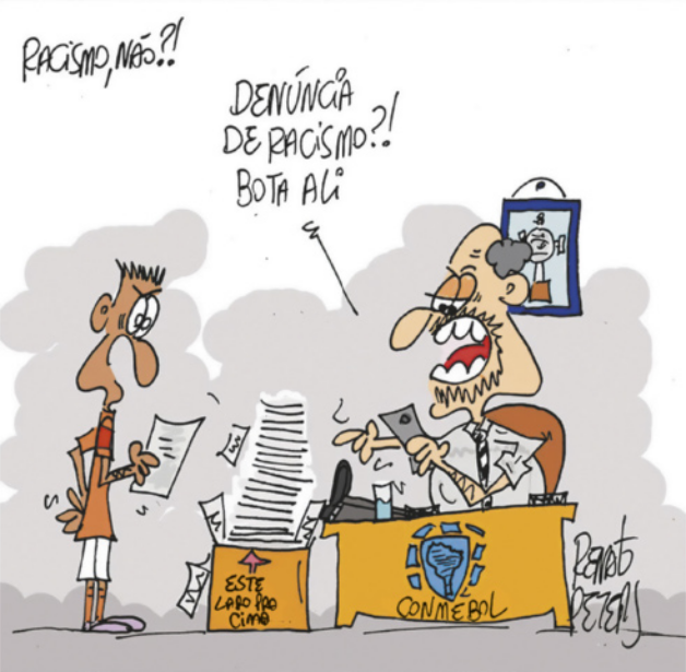 Charge intitulada 'Racismo, não?!': Um jogador de futebol segura uma carta diante de um homem sentado à mesa de um escritório, com o logotipo da Conmebol na mesa. Ele está com os pés apoiados sobre a mesa, segurando um smartphone, e diz ao jogador: 'Denúncia de racismo?! Bota ali', e aponta para uma caixa de papelão no chão, em que está escrito 'este lado para cima', contendo uma enorme pilha de papéis.