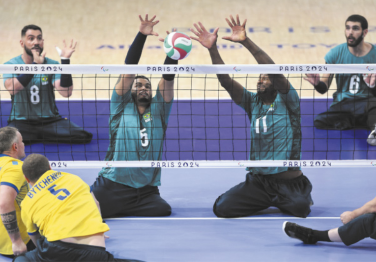 Fotografia de uma partida de vôlei sentado, em que dois jogadores da seleção brasileira, ajoelhados no chão, estendem os braços para o alto para bloquear a passagem da bola sobre a rede.