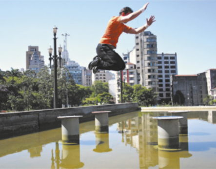Fotografia de Leonard Ribeiro Jacinto. Ele é um homem de cabelos curtos, de tênis, camiseta e calças flexíveis, saltando com os joelhos flexionados sobre um espelho d'água em uma praça.