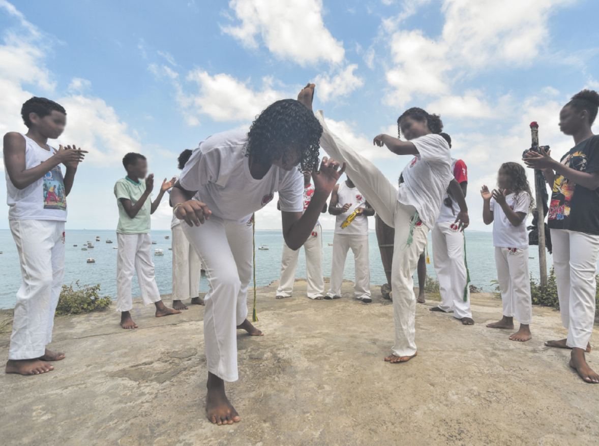Fotografia de um grupo de jovens jogando capoeira em roda. Ao centro estão duas garotas, uma delas chutando com a perna estendida no alto, e a outra se esquivando sob a perna da adversária, com o tronco inclinado para o lado. Os jovens em volta delas estão em pé, descalços e com uniformes brancos e calças de capoeira, batendo palmas, cantando e tocando instrumentos de percussão.