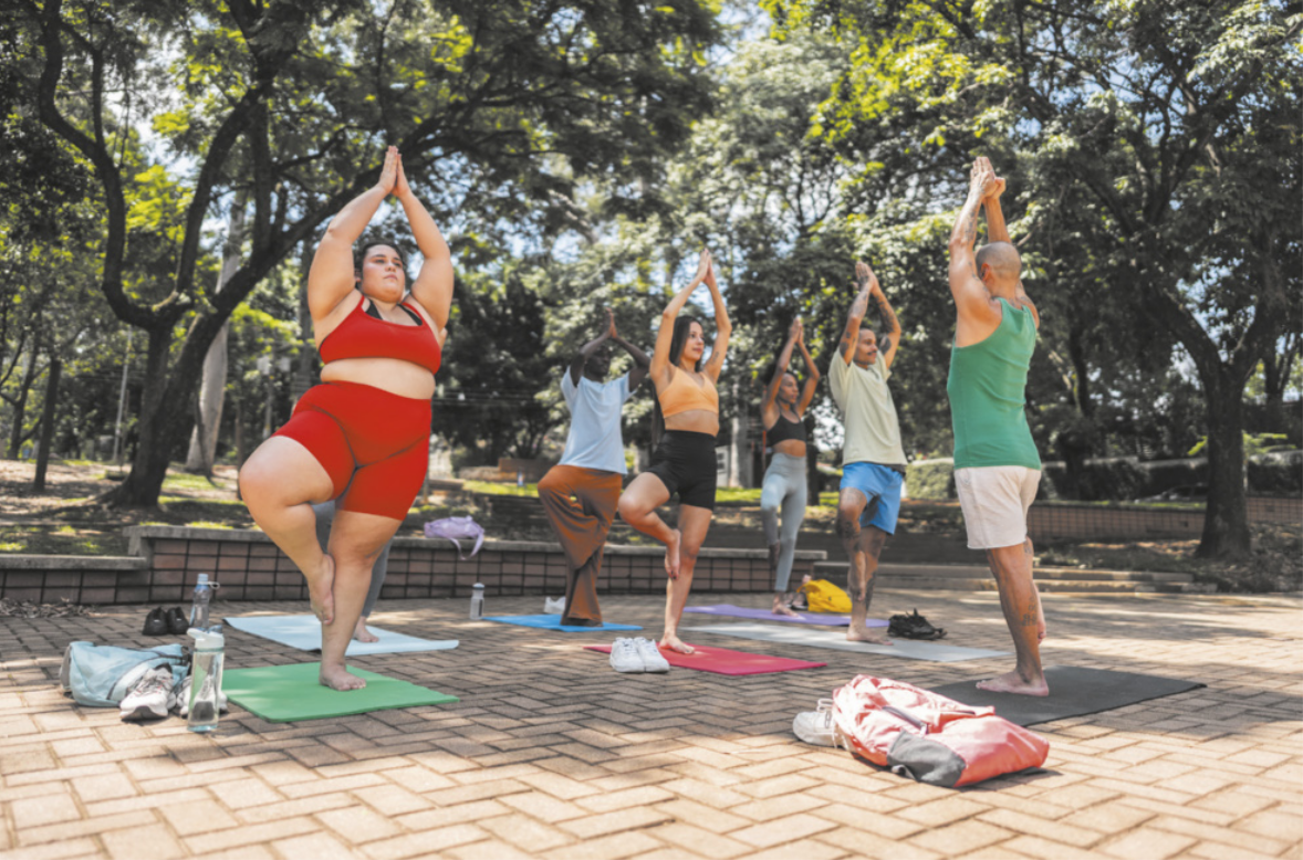 Fotografia de mulheres e homens em uma praça, praticando ioga sobre colchonetes estendidos no chão. Eles estão realizando uma postura apoiada sobre um pé só, apoiando a sola de um pé sobre a lateral interna da perna que está apoiada no chão, com a outra perna flexionada para o lado. Eles estão com o corpo ereto e os braços estendidos para o alto, unindo as palmas das mãos.