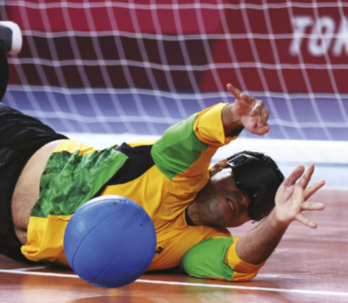 Fotografia de Romário Marques. Ele é um paratleta usando uma máscara sobre os olhos, deitado no chão com os braços estendidos em direção a uma bola, à frente de um gol em uma quadra de golbol.