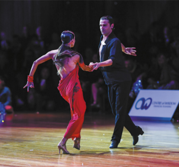 Fotografia de um casal dançando em um palco. O homem está de camisa aberta e segura a mão da mulher, inclinando o tronco para a frente e erguendo um dos braços para trás. A mulher, de vestido e salto alto, movimenta as pernas em direção ao parceiro, com os joelhos flexionados e um dos braços ao lado do corpo.