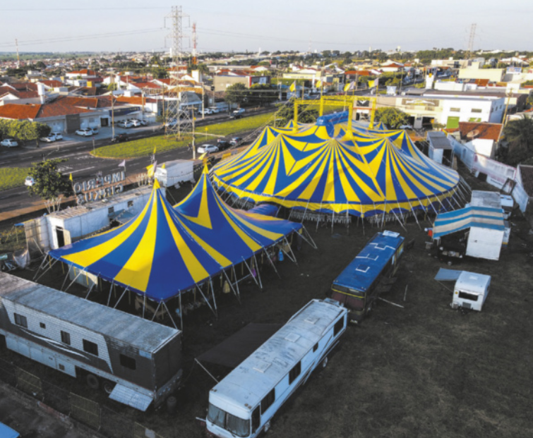 Fotografia aérea de um circo ao lado de uma via pública, em uma cidade com casas e edifícios baixos. O circo tem duas grandes tendas, e é circundado por ônibus e trailers.