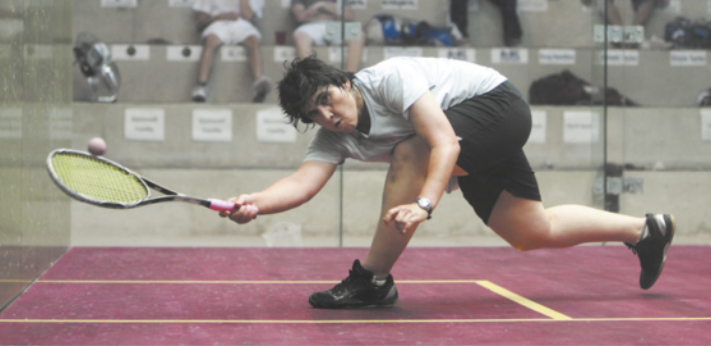Fotografia de Maria Toorpakai em uma quadra de squash, delimitada com paredes de vidro transparente e com uma arquibancada ao fundo. Ela é uma mulher com cabelos curtos, jogando squash com uma raquete na mão, agachando-se com as pernas afastadas e o tronco deitado, paralelo ao chão, estendendo a raquete para alcançar a bola.