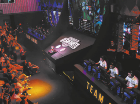 Fotografia de uma espécie de palco com duas mesas laterais com equipes jogando jogos eletrônicos diante de computadores. Ao centro do palco há um telão mostrando imagens do jogo, e diante dele está uma plateia de pessoas sentadas no chão, em pé e em cadeiras.