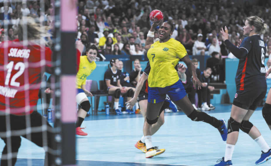 Fotografia de mulheres jogando handebol em uma quadra, com a plateia em uma arquibancada ao fundo. Uma das jogadoras veste a camiseta da seleção brasileira de handebol, e está saltando com a bola na mão erguida à altura da cabeça, em direção ao gol, defendido por uma goleira que está em pé, com as pernas afastadas e os joelhos flexionados, erguendo os braços.
