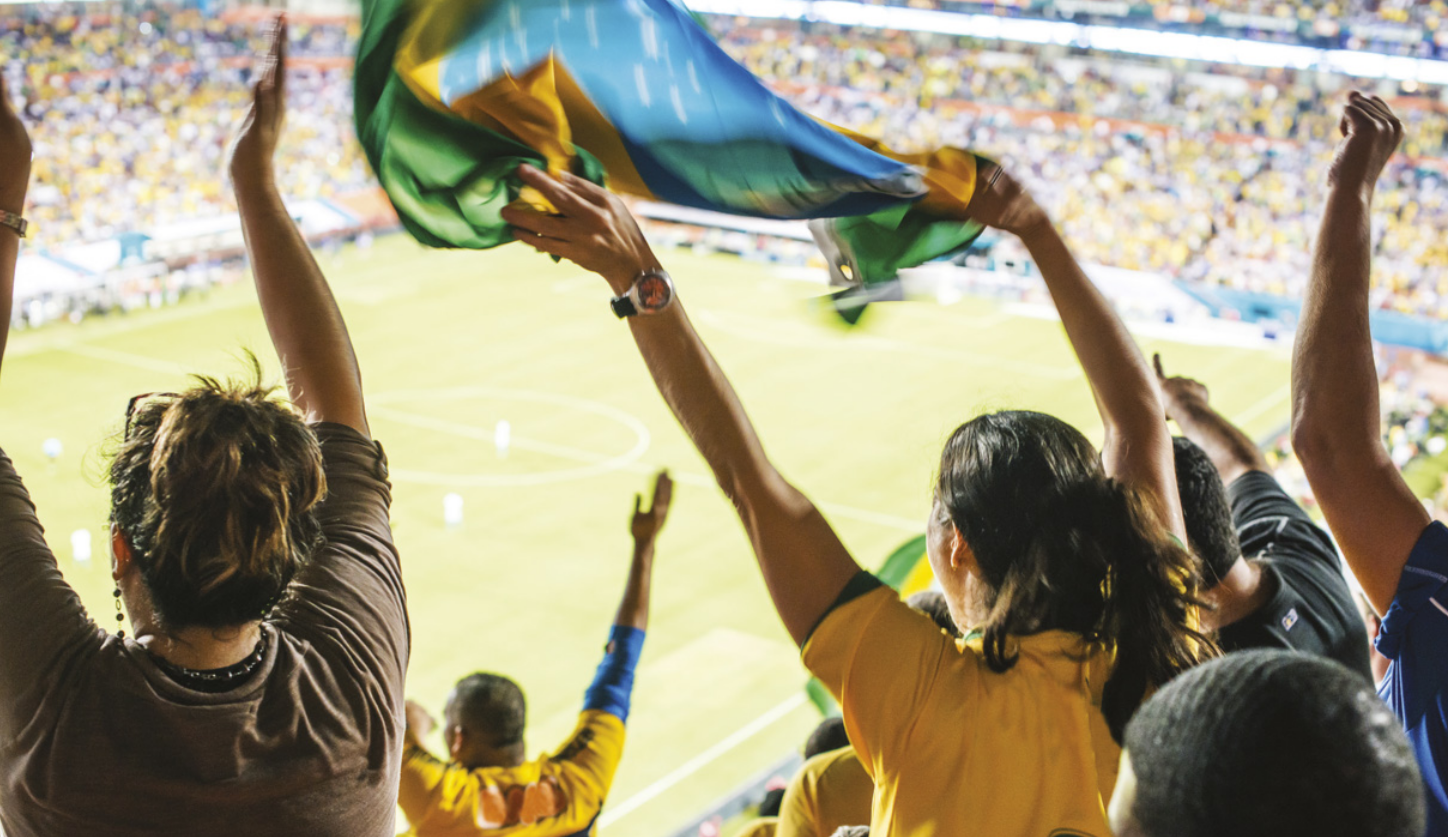 Fotografia de pessoas agitando uma bandeira do Brasil e erguendo os braços em uma arquibancada de estádio, observando de longe o campo de futebol.