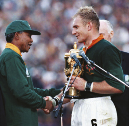 Fotografia de Nelson Mandela. Ele é um homem idoso de boné, cumprimentando com um aperto de mãos o atleta François Pienaar: um homem de cabelos louros curtos, segurando uma taça dourada.