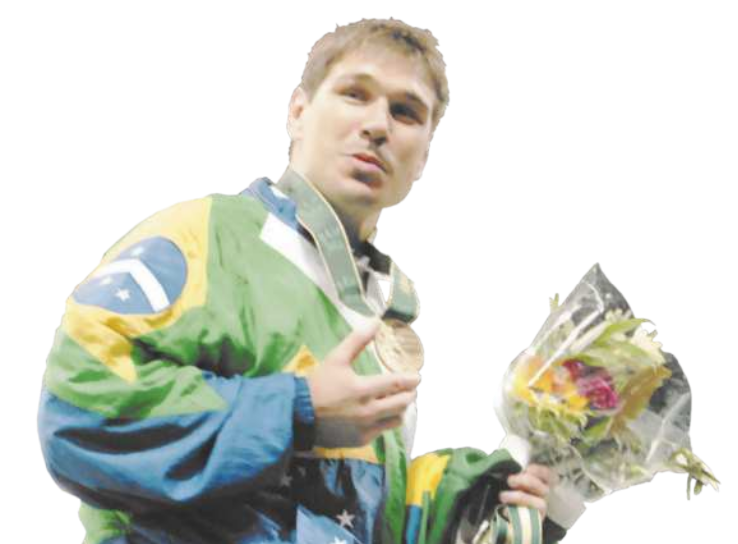 Fotografia de Aurélio Miguel. Ele é um homem de cabelos curtos, usando uma jaqueta com as cores da bandeira do Brasil, segurando uma medalha de bronze na mão, pendurada ao pescoço, e um buquê de flores na outra mão.