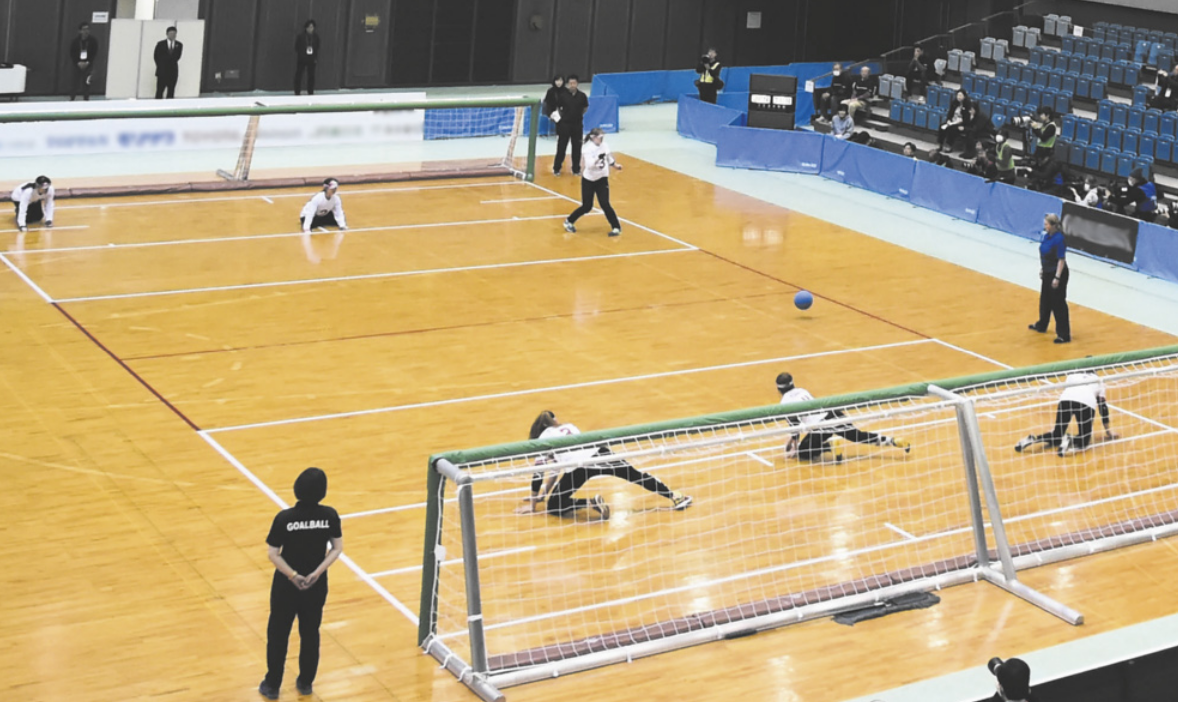 Fotografia de uma partida de golbol paralímpico, em uma quadra com piso de madeira. Duas equipes de três jogadoras com os olhos vendados defendem dois gols, que ocupam as linhas de fundo da quadra. Cinco jogadoras estão ajoelhadas no chão, e uma delas está em pé, diante da bola.