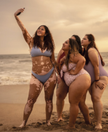 Fotografia de quatro mulheres de maiô e biquíni em uma praia, posando para uma selfie feita por uma delas, que está à frente, segurando o smartphone no alto, focalizando o grupo.