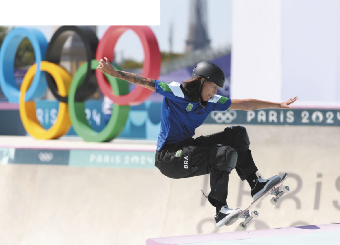 Fotografia de Isadora Pacheco. Ela é uma garota com cabelos longos e lisos, andando de skate com um capacete e joelheiras. Ela está com os braços abertos e estendidos ao lado do corpo, realizando uma manobra com o skate no ar, no topo de uma rampa em uma pista de skate olímpica.