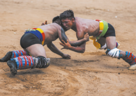 Fotografia de dois homens indígenas lutando sobre a terra, próximos ao chão, agarrando-se mutuamente com os braços. Eles estão com os corpos inclinados para a frente, quase deitados no ar, apoiados sobre as pernas flexionadas, tentando derrubar o oponente.