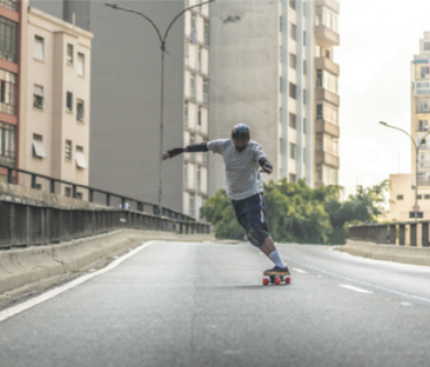 Fotografia de um homem andando de skate sobre o asfalto de um viaduto para carros, usando capacete, luvas, joelheiras e cotoveleiras, com prédios ao fundo.