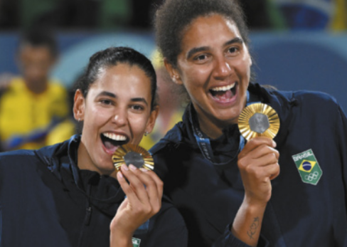 Fotografia de Duda e Ana Patrícia. Elas são duas mulheres com os cabelos presos, sorrindo e segurando medalhas de ouro penduradas ao pescoço.